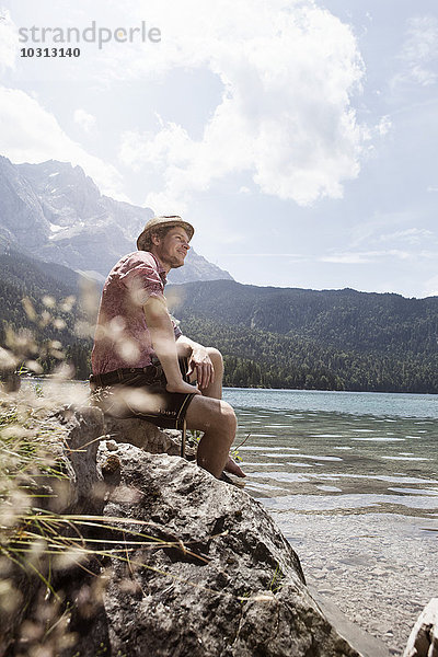 Deutschland  Bayern  Eibsee  lächelnder Mann in Lederhose sitzend am Seeufer