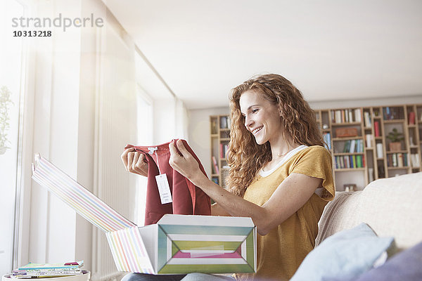 Lächelnde Frau zu Hause auf der Couch sitzend  Paket mit Kleidungsstück auspackend