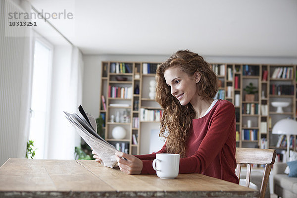 Lächelnde Frau zu Hause sitzend am Holztisch mit Tasse Zeitung lesen
