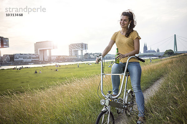 Deutschland  Köln  junge Frau mit Fahrrad auf der Wiese am Rhein bei Abenddämmerung