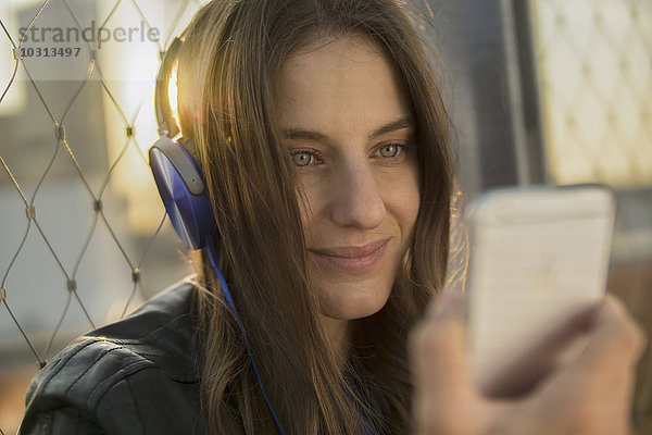 Deutschland  Frankfurt  Porträt einer lächelnden Frau  die Musik mit Kopfhörern hört und auf das Smartphone schaut.
