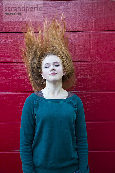Porträt eines jungen Mädchens  das seine Haare vor einer roten Holzwand wirft.