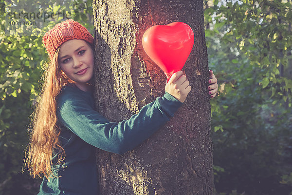 Porträt eines lächelnden Teenagermädchens mit herzförmigem Ballon an einem Baum