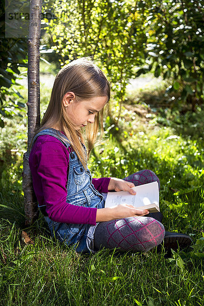 Kleines Mädchen sitzt auf einer Wiese im Garten und liest ein Buch.