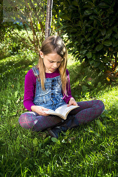 Kleines Mädchen sitzt auf einer Wiese im Garten und liest ein Buch.