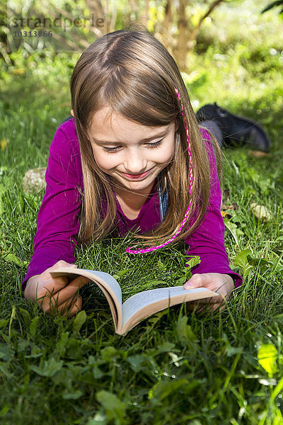 Glückliches kleines Mädchen liegt auf einer Wiese im Garten und liest ein Buch.