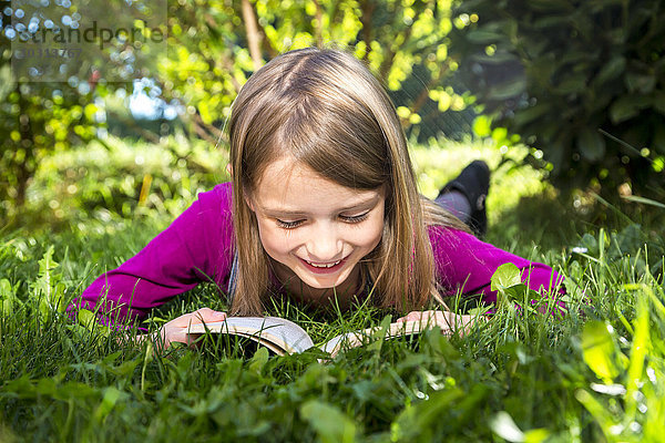 Kleines Mädchen liegt auf einer Wiese im Garten und liest ein Buch.