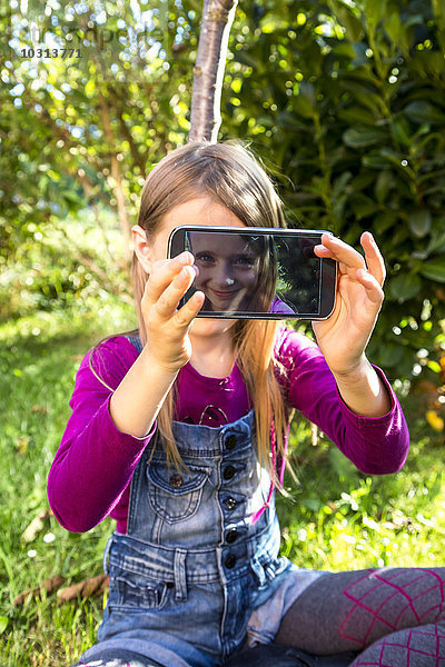 Kleines Mädchen sitzt auf einer Wiese im Garten und zeigt das Display eines Smartphones mit ihrem Selfie.
