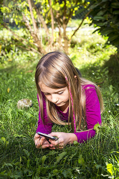 Kleines Mädchen liegt auf einer Wiese im Garten und schaut auf das Smartphone.