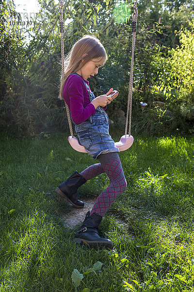 Kleines Mädchen sitzt auf einer Schaukel im Garten und schaut auf das Smartphone.