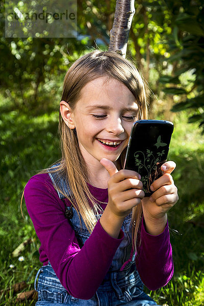 Fröhliches kleines Mädchen beim Blick auf das Smartphone im Garten