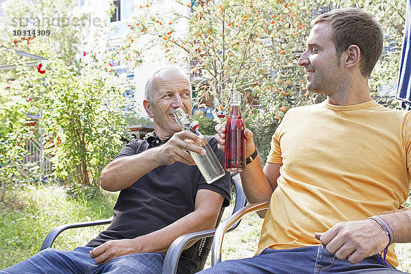 Vater und sein erwachsener Sohn sitzen im Garten und stoßen mit Getränken an.
