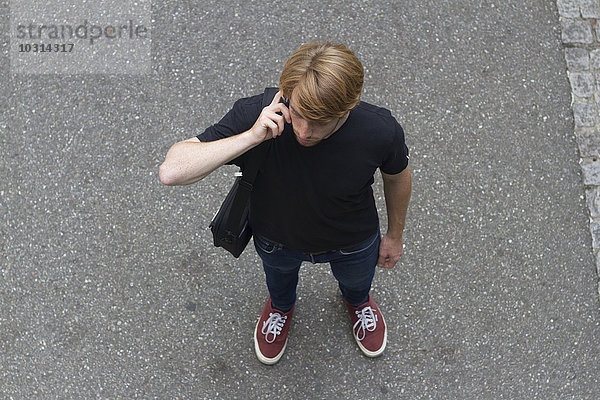 Portrait eines jungen Mannes  der auf einer Straße steht und mit einem Smartphone telefoniert.
