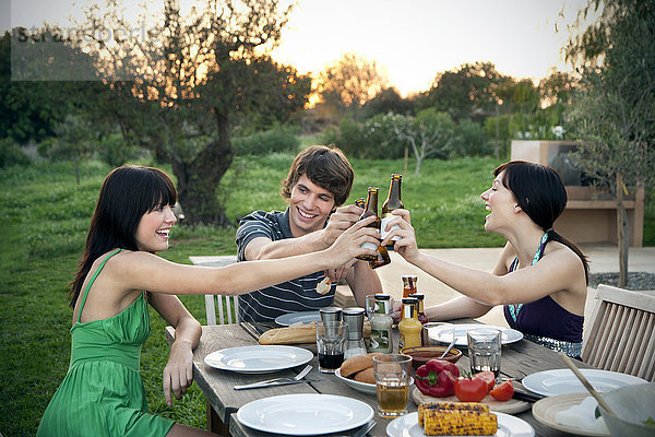 Glückliche Freunde sitzen am Gartentisch und grillen Bierflaschen.