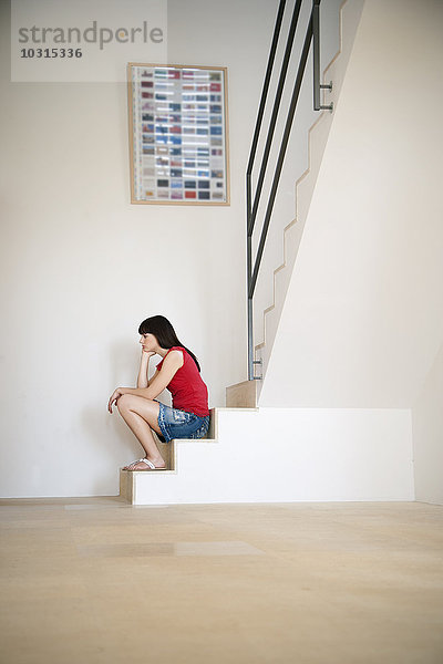 Nachdenkliche junge Frau sitzt auf einer Treppe in einem hellen Raum.