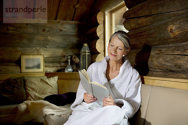 Lächelnde Seniorin sitzt auf einer Bank im Bademantel und liest ein Buch.