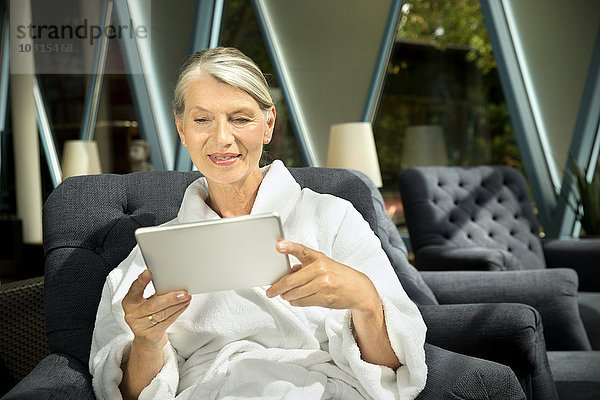 Seniorin im Bademantel sitzend im Sessel mit digitalem Tablett