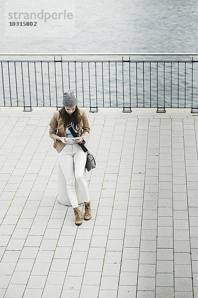 Junge Frau sitzt auf einem Poller an der Promenade und orientiert sich mit einem digitalen Tablett.