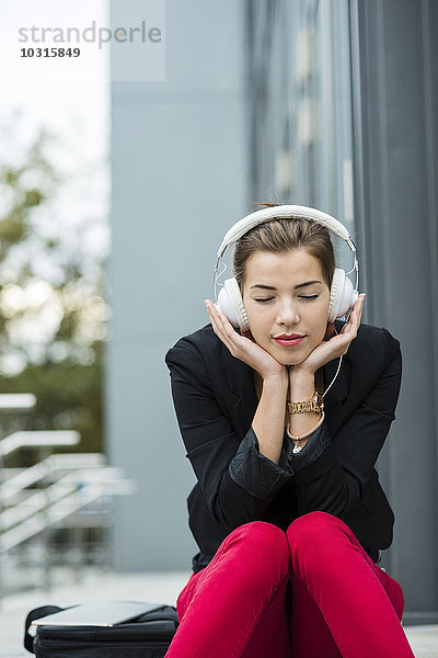 Junge Frau mit geschlossenen Augen sitzt auf einer Treppe und hört Musik über Kopfhörer.