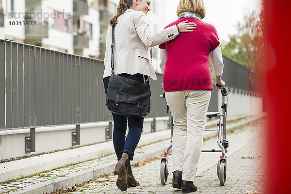 Erwachsene Enkelin  die ihrer Großmutter beim Laufen mit Rollator hilft  Rückansicht