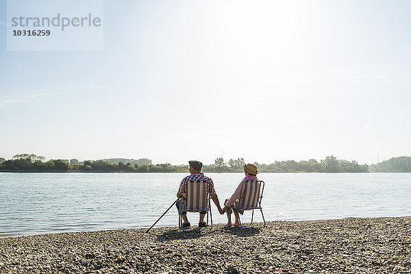 Deutschland  Ludwigshafen  Rückansicht eines älteren Ehepaares  das Hand in Hand auf Klappstühlen am Flussufer sitzt