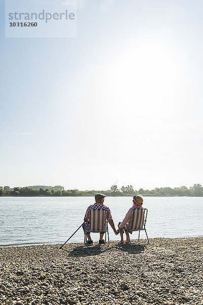Deutschland  Ludwigshafen  Rückansicht eines älteren Ehepaares  das Hand in Hand auf Klappstühlen am Flussufer sitzt