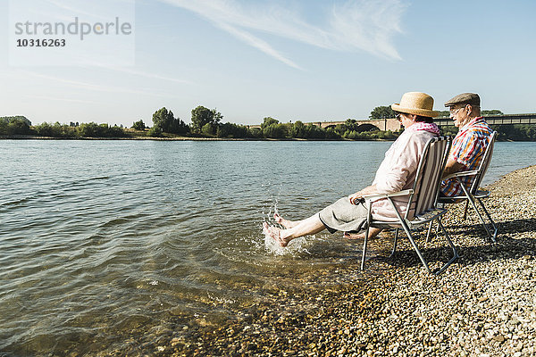Deutschland  Ludwigshafen  Seniorenpaar sitzt auf Klappstühlen am Flussufer und spritzt mit den Füßen im Wasser.