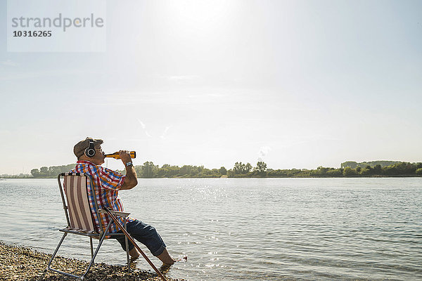 Deutschland  Ludwigshafen  Senior mit Kopfhörer auf Klappstuhl am Flussufer beim Biertrinken