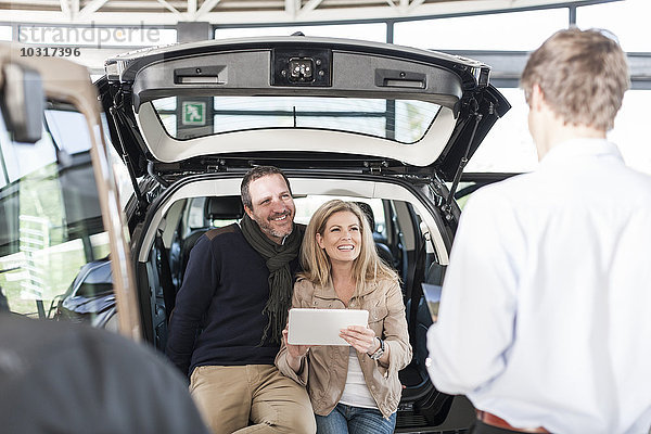 Lächelndes Paar sitzt im Kofferraum des neuen Autos im Autohaus