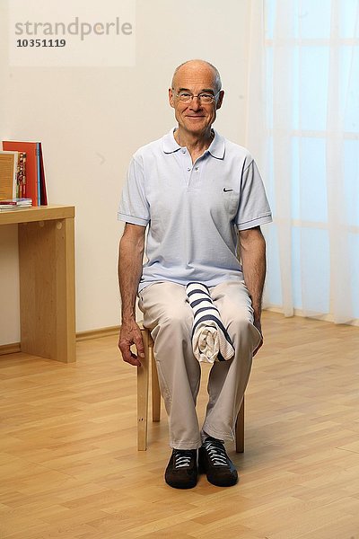 Älterer Mann sitzt auf einem Stuhl und macht Knieübungen - Senior macht Gymnastik - weniger Schmerzen