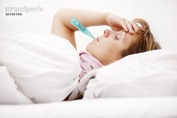 Junge erwachsene Frau  die krank zu Hause im Bett liegt und Fieber hat  misst die Körpertemperatur mit einem digitalen Fieberthermometer
