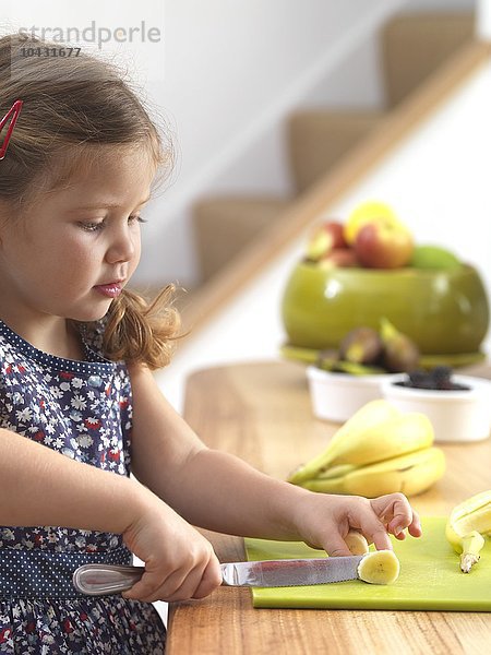 MODELL FREIGEGEBEN. Junges Mädchen schneidet eine Banane. Drei Jahre altes Mädchen bereitet sich einen Snack.
