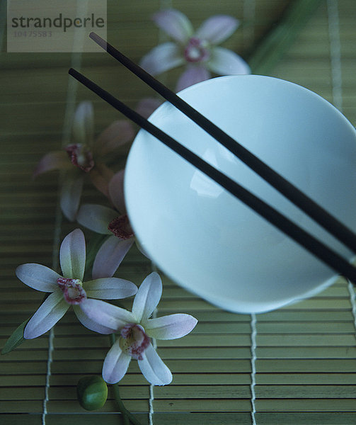 Essstäbchen und eine Reisschale auf einer Bambusmatte mit verstreuten Orchideenblütenknospen. Essstäbchen sind Esswerkzeuge  die in einer Hand gehalten werden und im Fernen Osten verwendet werden.