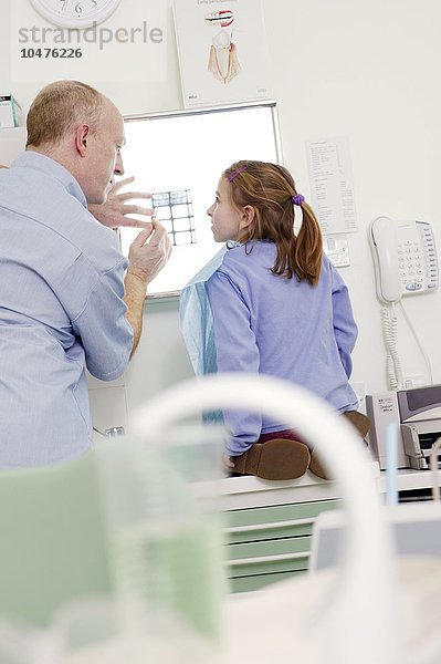 MODELL FREIGEGEBEN. Zahnärztliches Röntgen. Ein Mädchen lässt sich von einem Zahnarzt das Röntgenbild ihres Mundes erklären. Der Zahnarzt hält das Röntgenbild gegen einen Leuchtkasten  während er Teile ihres Mundes zeigt Zahnröntgenbild