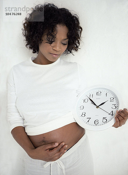 MODELL FREIGEGEBEN. Zeitangaben in der Schwangerschaft. Schwangere Frau  die in einer Hand eine Uhr hält  während sie mit der anderen Hand ihren geschwollenen Bauch stützt. Sie ist in der 30. Woche schwanger. Die Uhr könnte die Zeitspanne darstellen  in der sich ein Baby entwickelt (die Schwangerschaftszeit). Eine Vollschwangerschaft dauert zwischen 37 und 42 Wochen  also etwa neun Monate. Wenn das Baby einige Wochen zu früh kommt  wird es als Frühgeburt bezeichnet. Wenn es zu spät kommt  können die Wehen eingeleitet werden. Schwangerschaftszeiten