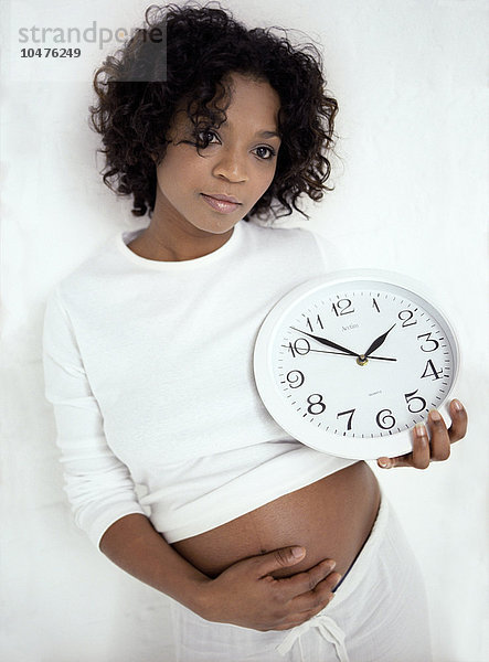 MODELL FREIGEGEBEN. Zeitangaben in der Schwangerschaft. Schwangere Frau  die in einer Hand eine Uhr hält  während sie mit der anderen Hand ihren geschwollenen Bauch stützt. Sie ist in der 30. Woche schwanger. Die Uhr könnte die Zeitspanne darstellen  in der sich ein Baby entwickelt (die Schwangerschaftszeit). Eine Vollschwangerschaft dauert zwischen 37 und 42 Wochen  also etwa neun Monate. Wenn das Baby einige Wochen zu früh kommt  wird es als Frühgeburt bezeichnet. Wenn es zu spät kommt  können die Wehen eingeleitet werden. Schwangerschaftszeiten