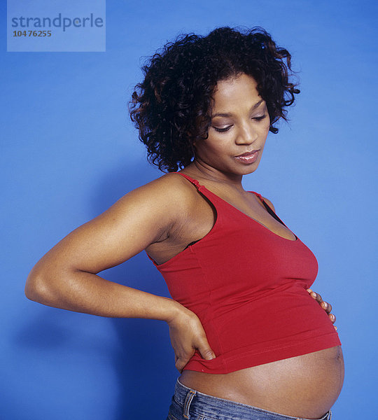 MODELL FREIGEGEBEN. Eine schwangere Frau hält sich den schmerzenden Rücken  da das Gewicht des ungeborenen Kindes ihre Rückenmuskulatur belastet. Sie ist in der 30. Woche schwanger. Eine volle Schwangerschaft dauert zwischen 37 und 42 Wochen. Schwangerschaft Rückenschmerzen