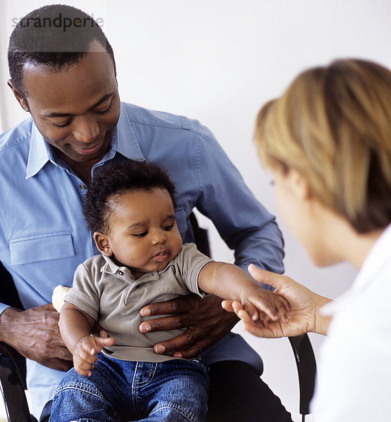 MODELL FREIGEGEBEN. Pädiatrische Untersuchung. Arzt untersucht einen 5 Monate alten Jungen  der auf dem Schoß seines Vaters sitzt Pädiatrische Untersuchung