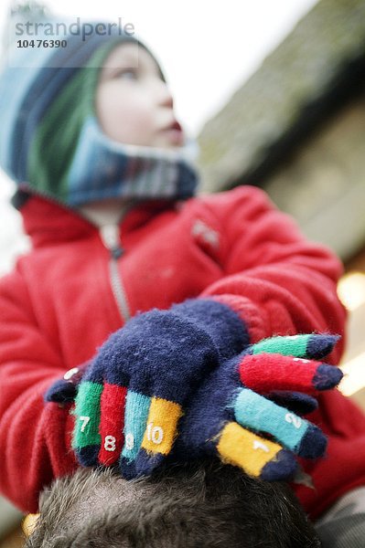 MODELL FREIGEGEBEN. Kind mit Handschuhen. Ein Kind sitzt auf den Schultern eines Erwachsenen und trägt Winterhandschuhe. Die Finger des Handschuhs sind unterschiedlich gefärbt und mit den Zahlen 1-10 bedruckt  um dem Kind das Zählen zu erleichtern. Kind mit Handschuhen