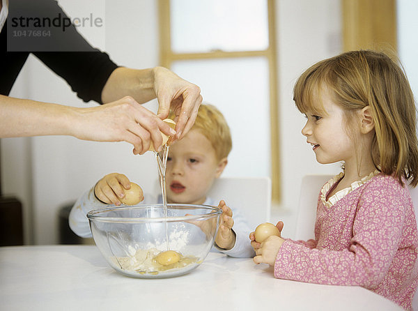 MODELL FREIGEGEBEN. Kuchen backen. Zwei dreijährige Kinder sehen ihrer Mutter zu  wie sie ein Ei in eine Rührschüssel aufschlägt. Kuchen backen