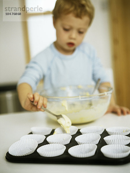 MODELL FREIGEGEBEN. Kuchen backen. Dreijähriger Junge  der den Kuchenteig in einzelne Papierförmchen gibt  bevor sie im Ofen gebacken werden Kuchen backen