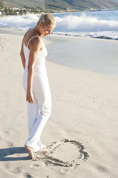 MODELL FREIGEGEBEN. Herz im Sand. Frau zeichnet eine Herzform an einem Sandstrand Herz im Sand