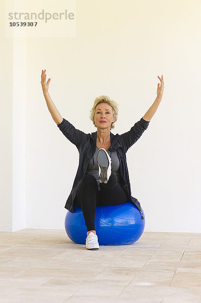 Reife Frau sitzt auf einem Fitnessball mit erhobenen Armen und einem Bein nach oben.