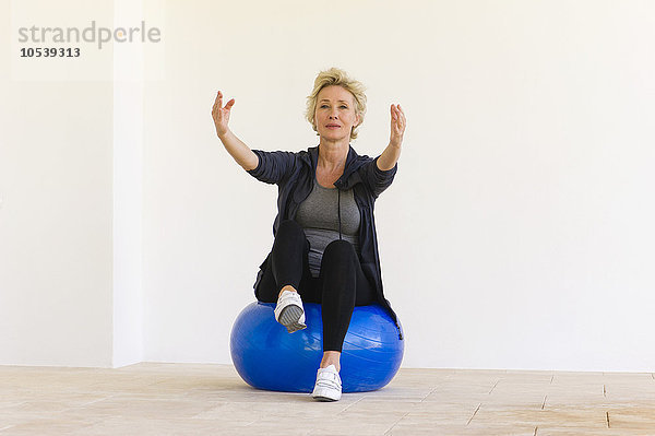 Reife Frau sitzt auf einem Fitnessball mit ausgestreckten Armen und einem Bein nach oben