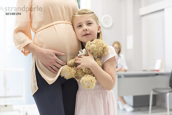 Portrait Mädchen mit Teddybär umarmt schwangere Mutter in Arztpraxis