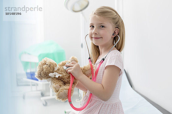 Portrait lächelndes Mädchen mit Stethoskop auf Teddybär im Untersuchungszimmer