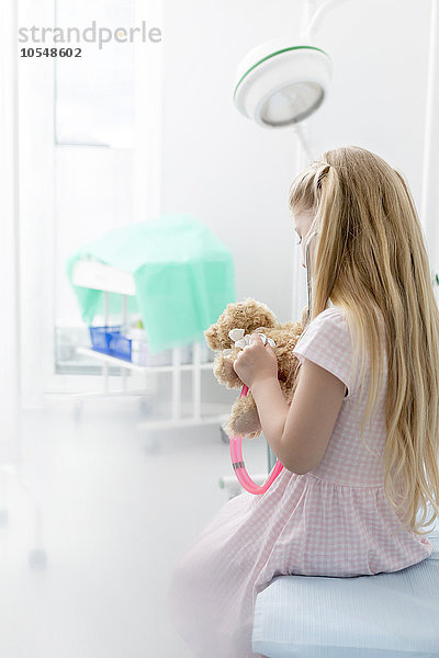 Mädchen mit Stethoskop auf Teddybär im Untersuchungszimmer