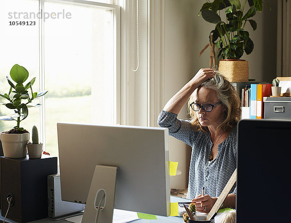 Gestresste Frau mit Hand im Haar hält Kreditkarte am Computer im Home-Office