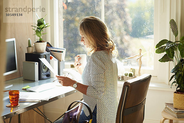 Frau trinkt Kaffee und liest Papierkram am Schreibtisch im sonnigen Home-Office.