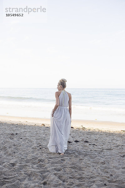 Blonde Frau in einem langen Kleid  die an einem Sandstrand steht.